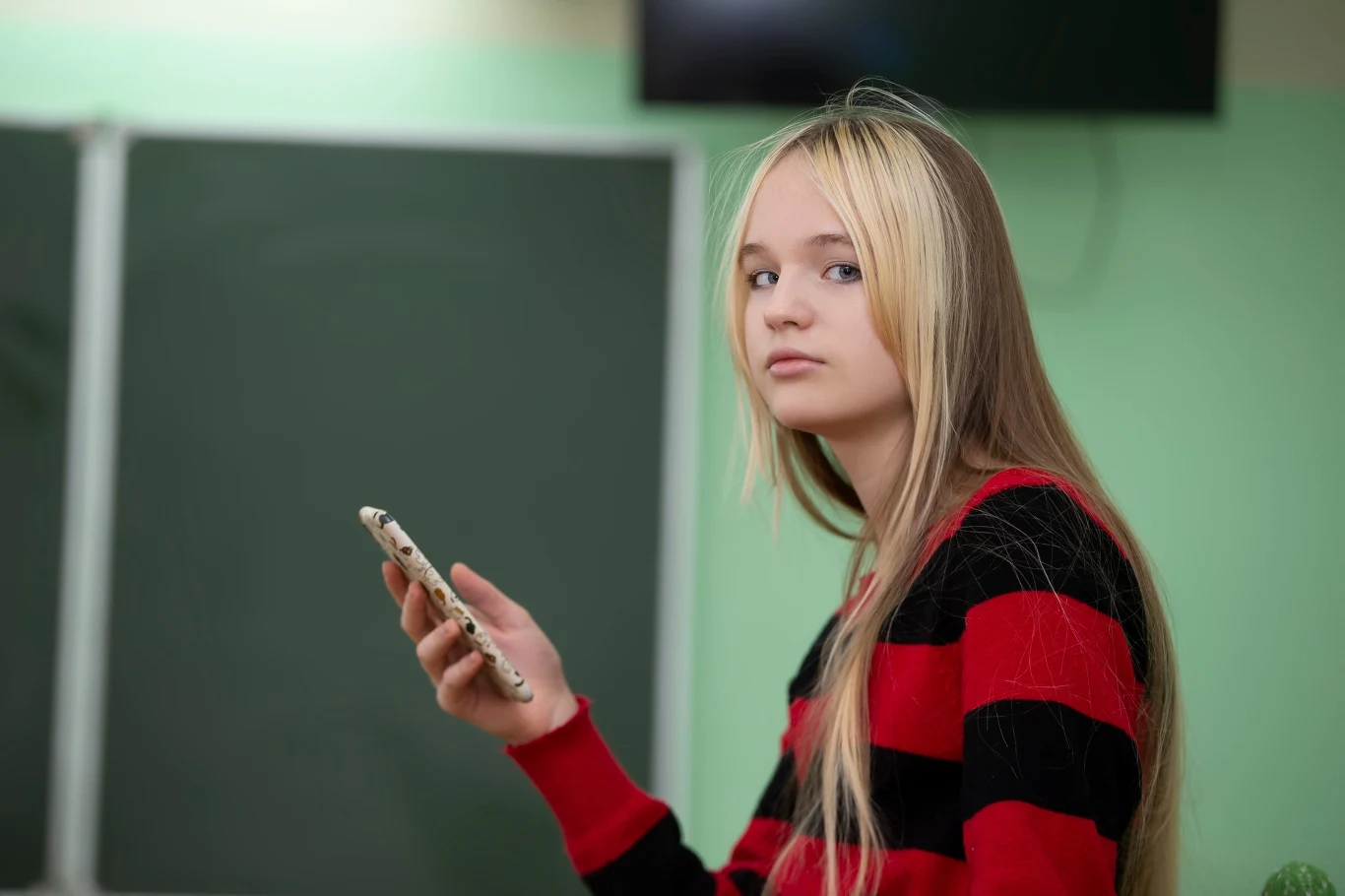 Nastolatka z długimi blond włosami ubrana w sweter w czerwono-czarne paski siedzi w klasie i trzyma w ręku smartfon, patrząc lekko w bok.