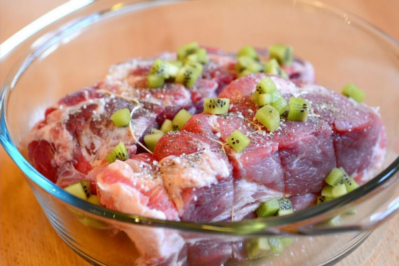 Marynata z kiwi skutecznie zmiękczy mięso Kawałki surowego mięsa w szklanej misce, posypane kostkami kiwi i przyprawami, przygotowane do marynowania.