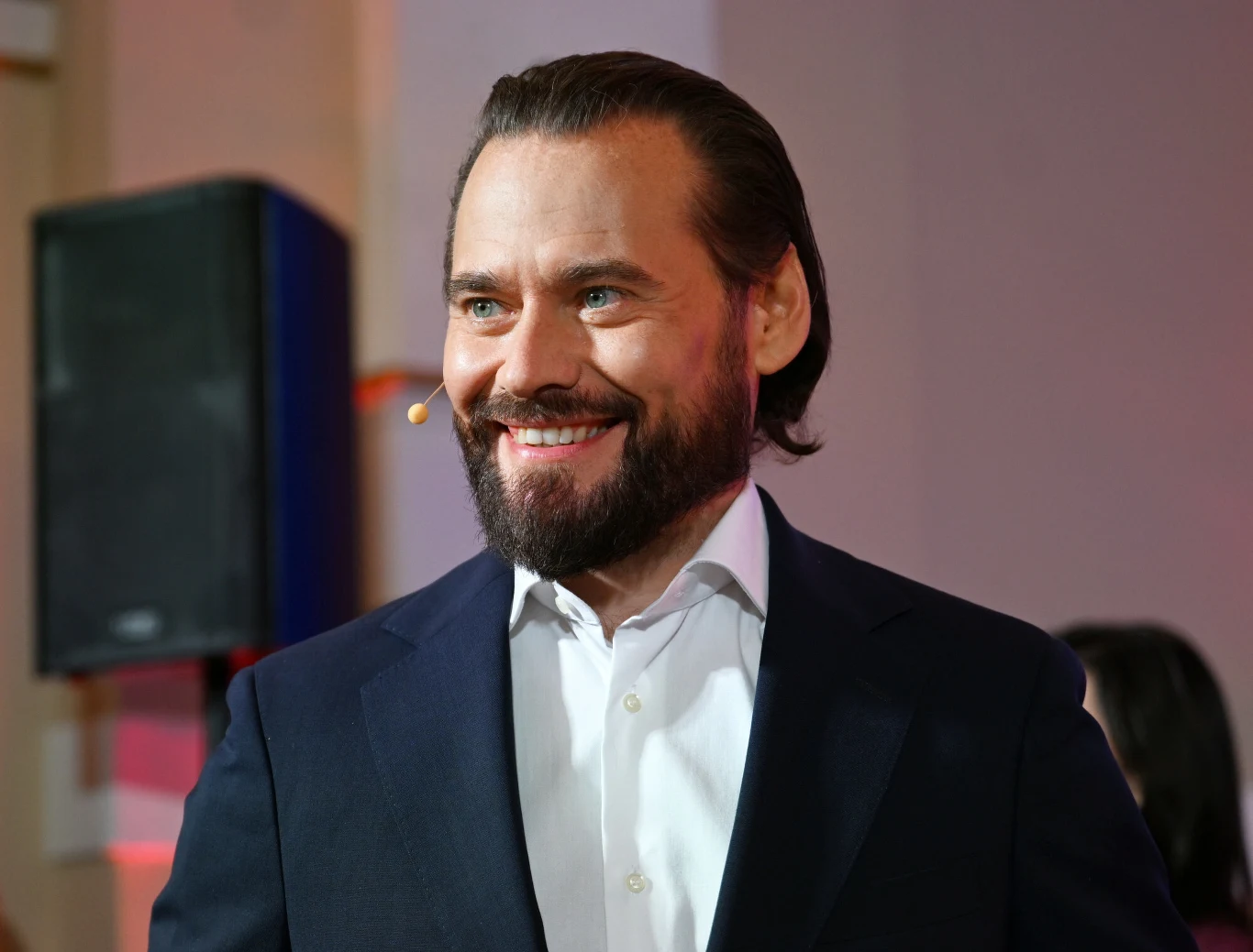 Krzysztof Stanowski Uśmiechnięty mężczyzna w garniturze i białej koszuli, z mikrofonem na uchu, stoi w jasnym pomieszczeniu o eleganckim charakterze.