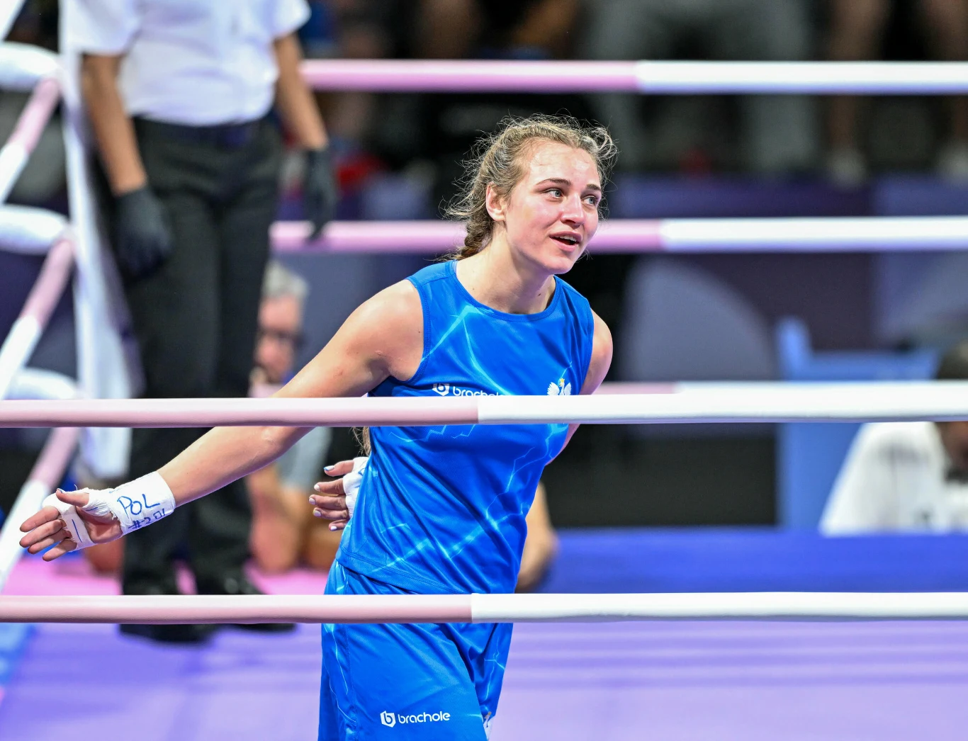 bokserka w niebieskim stroju sportowym stoi na ringu, z owiniętymi taśmami rękami, wygląda na zmęczoną, ale zadowoloną, w tle fragment sylwetki sędzi i linie ringu
