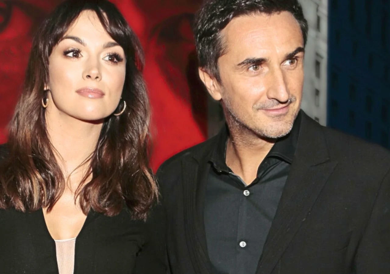 Paulina Krupińska i Sebastian Karpiel-Bułecka Kobieta z długimi, ciemnymi włosami i mężczyzna o ciemnych włosach stoją obok siebie w eleganckich, czarnych ubraniach, na tle czerwono-czarnej grafiki.
