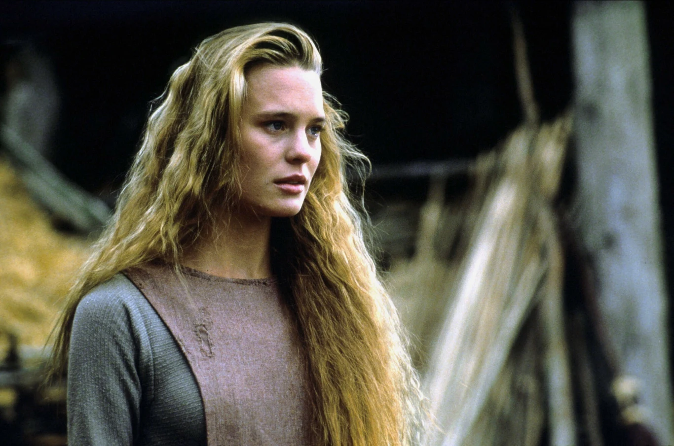 młoda kobieta o długich, falowanych blond włosach w prostej, średniowiecznej sukni, stojąca na tle wiejskiej zabudowy z drewnianych bali