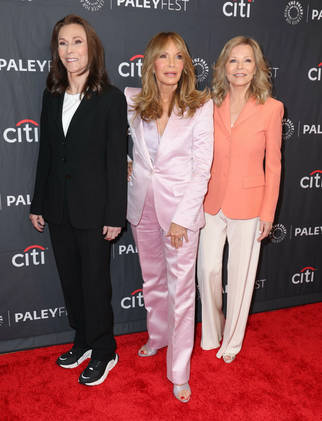 Trzy kobiety w eleganckich garniturach stoją na czerwonym dywanie na tle ściany z logotypami PaleyFest i Citi; jedna ma na sobie czarny garnitur i sportowe buty, druga jasnoróżowy garnitur i szpilki, trzecia łososiowy żakiet i białe spodnie.