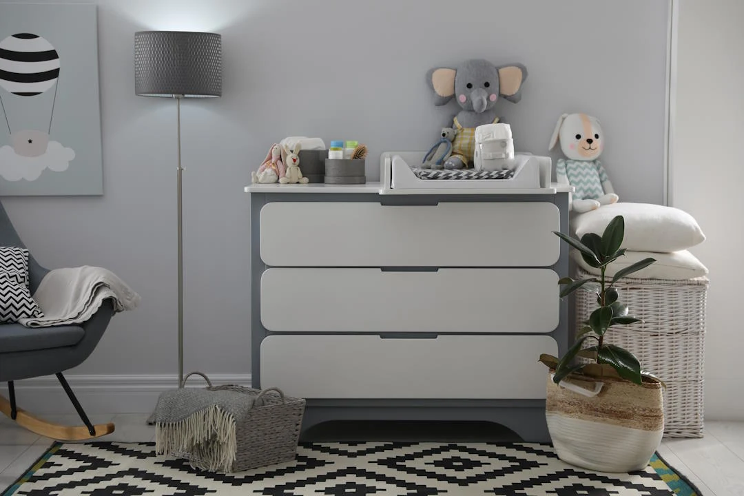 komoda z przewijakiem ustawiona na geometrycznym dywanie, na komodzie pluszowy słoń, zabawkowy pies, organizer i kosmetyki dziecięce; obok komody kosz z rośliną doniczkową i kosz na pranie, w tle szara lampa stojąca oraz obraz z motywem balonu