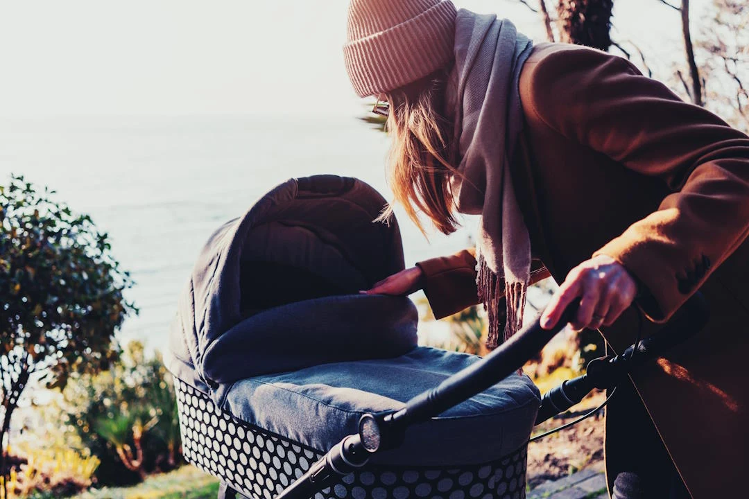 kobieta w płaszczu i czapce pochyla się nad wózkiem dziecięcym, otoczona jesienną aurą nad brzegiem wody
