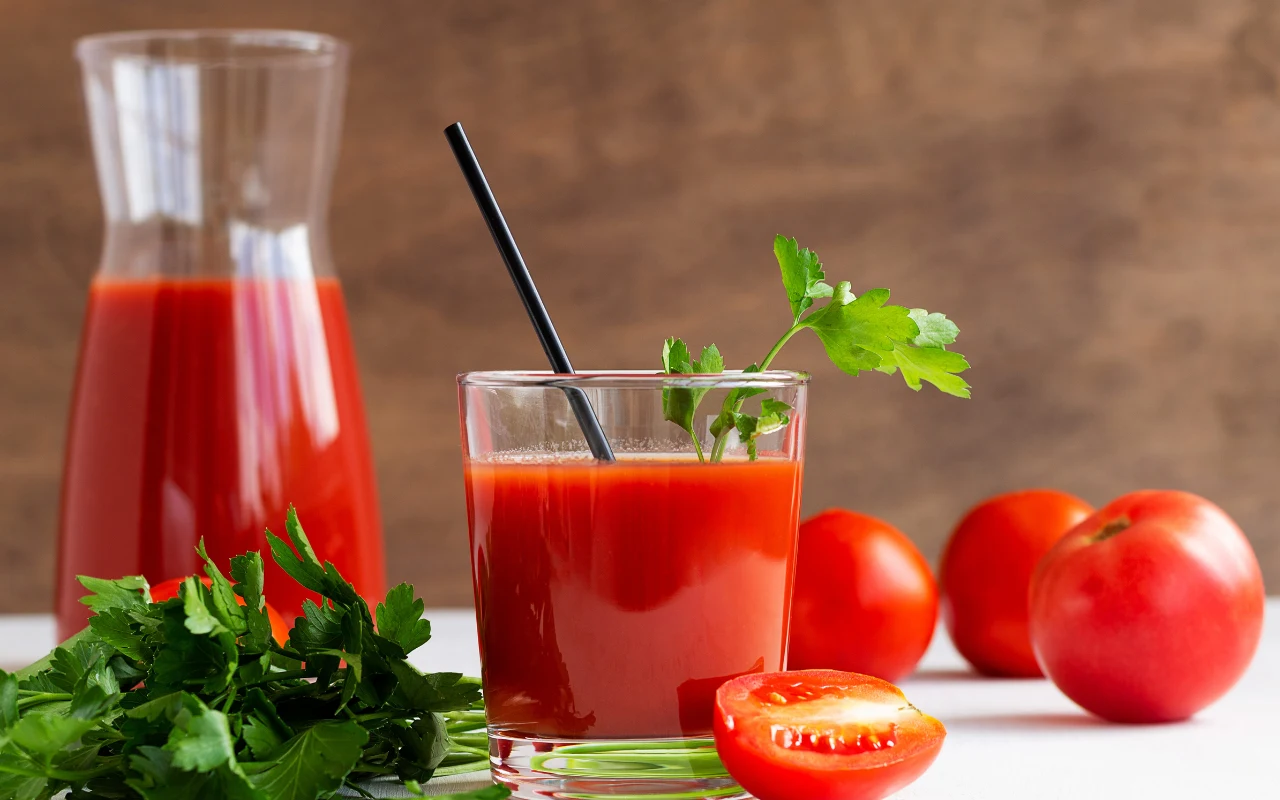 Szklanka soku pomidorowego z czarną słomką i gałązką natki pietruszki, obok świeża natka, karafka z sokiem oraz kilka dojrzałych pomidorów na jasnej powierzchni.