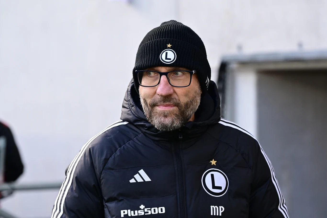 Mężczyzna w czarnej kurtce sportowej oraz czapce z emblematem Legii Warszawa, z brodą i okularami, stoi na tle jasnej ściany.