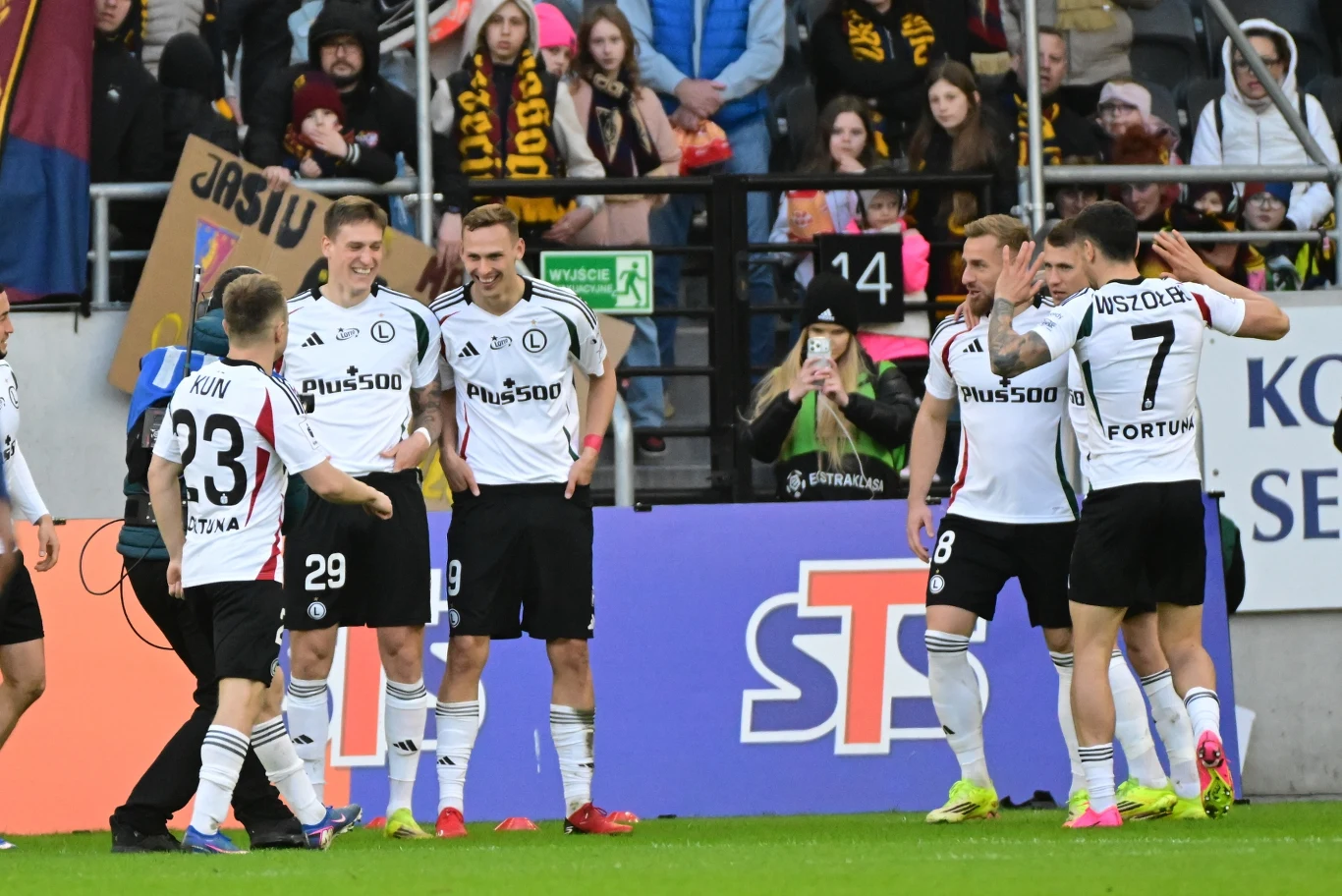 piłkarze w biało-czarnych strojach Legii Warszawa świętują zdobycie bramki na murawie stadionu, za nimi trybuny wypełnione kibicami