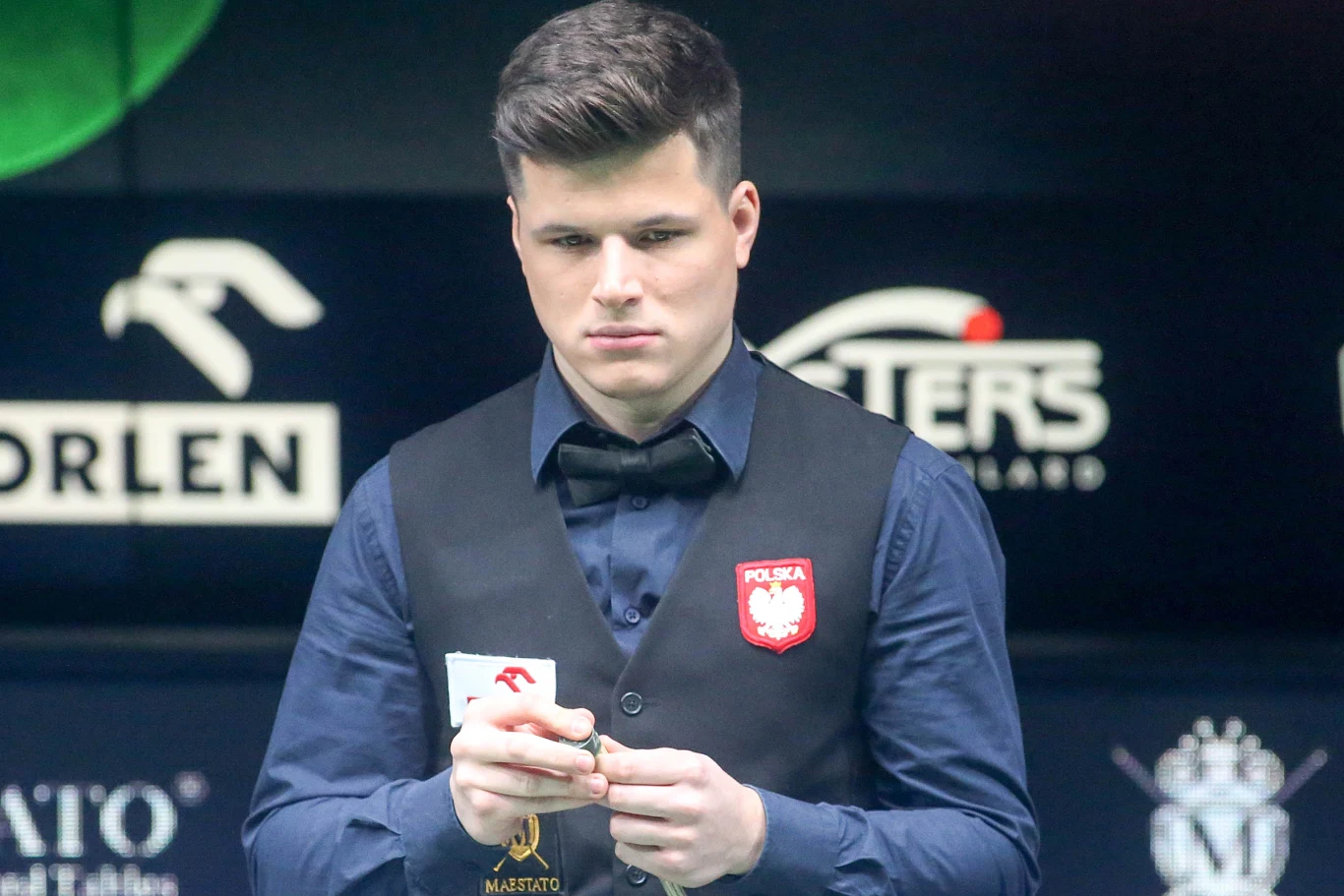 młody mężczyzna w eleganckim stroju bilardowym z godłem Polski na piersi trzyma kredę do kija, patrzy skoncentrowanym wzrokiem, w tle widoczne są logotypy sponsorów