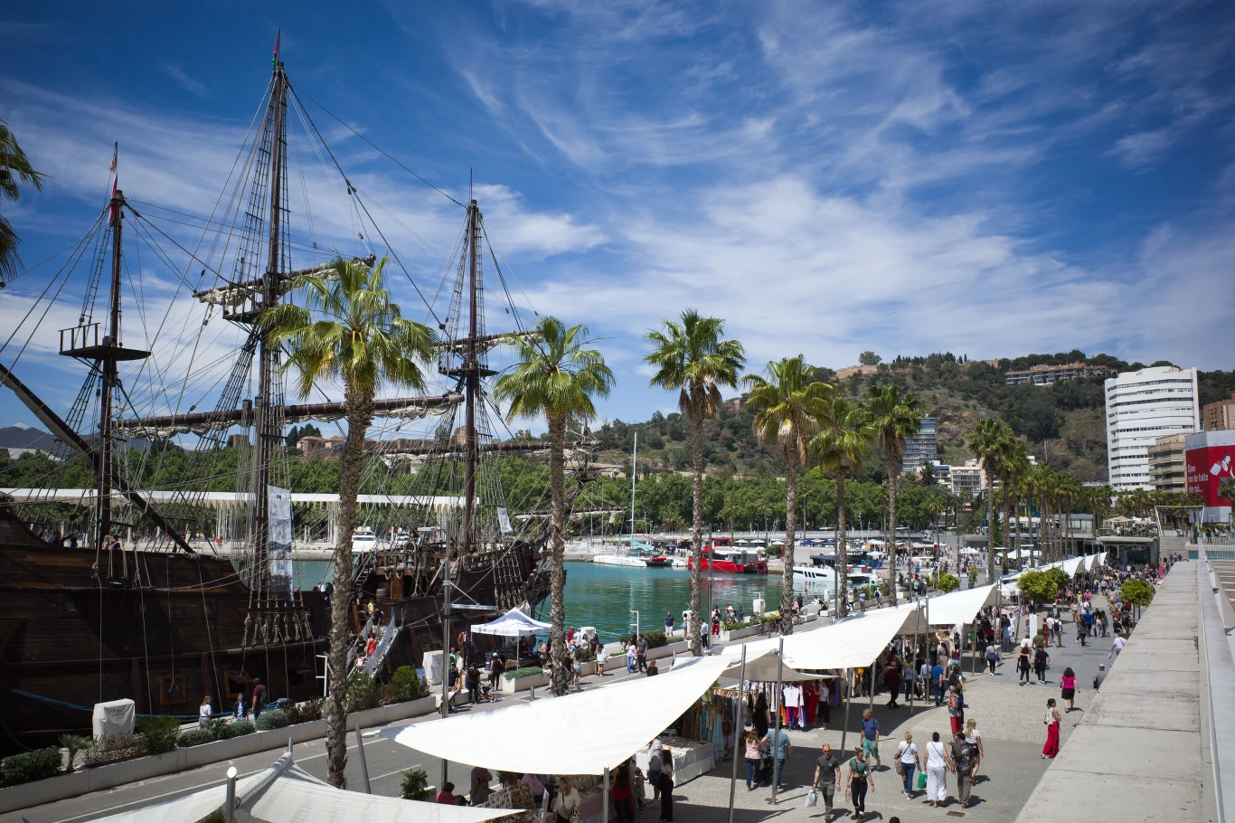 duży żaglowiec cumujący przy palmowym deptaku, obok którego rozstawione są stoiska i przechadzają się ludzie, w tle marinę wypełniają jachty i zielone wzgórza, a nad całością rozciąga się błękitne niebo