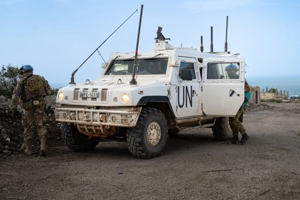 Biały opancerzony pojazd ONZ z napisem „UN” i żołnierzami w niebieskich hełmach na tle skalistego terenu.