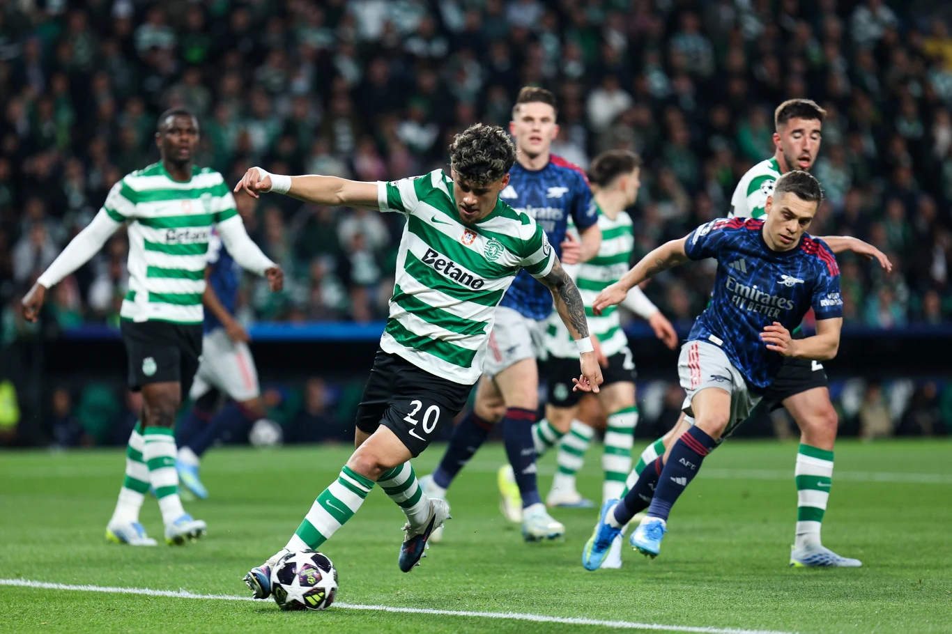 zawodnik w zielono-białej koszulce oddaje strzał na bramkę, a wokół niego kilku piłkarzy rywalizujących drużyn próbuje interweniować podczas meczu piłkarskiego na stadionie pełnym kibiców