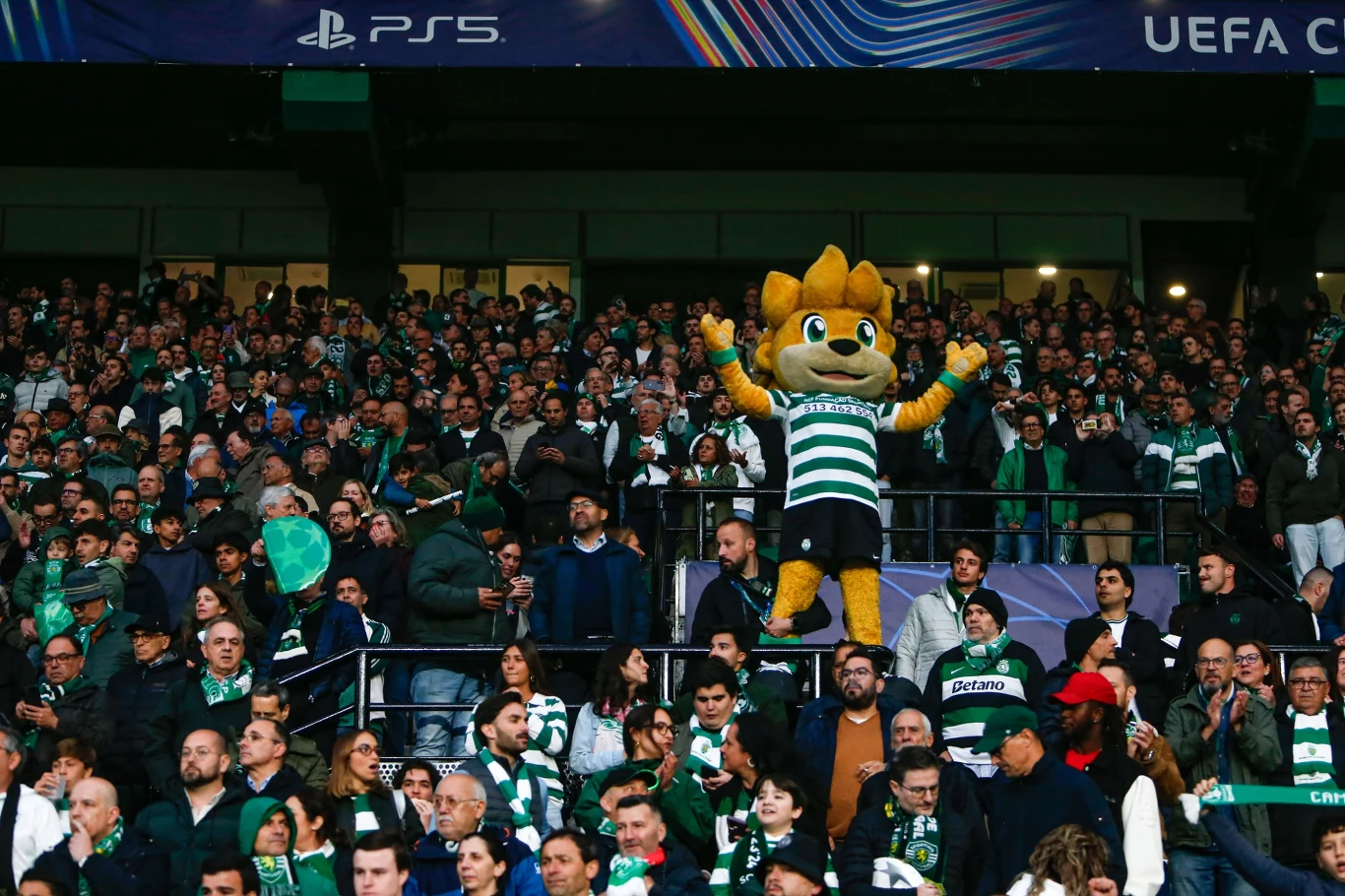 maskotka w zielono-białej koszulce piłkarskiej unosi łapy w tłumie kibiców na trybunach podczas meczu UEFA Champions League, wokół liczne osoby ubrane w klubowe barwy
