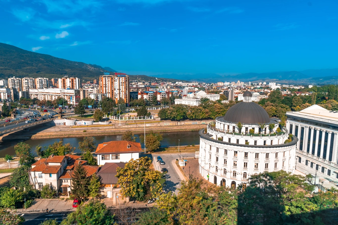 dominująca okrągła biała budowla z kopułą otoczona zielenią w centrum miasta, szeroka rzeka przecinająca krajobraz, w tle nowoczesne wieżowce i pasmo górskie pod błękitnym niebem