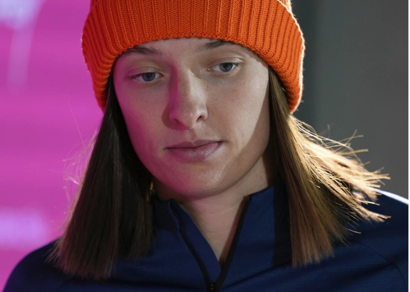 młoda kobieta w pomarańczowej czapce i granatowej kurtce patrząca w dół, o poważnym wyrazie twarzy, z rozpuszczonymi włosami na tle rozmytego różowego tła