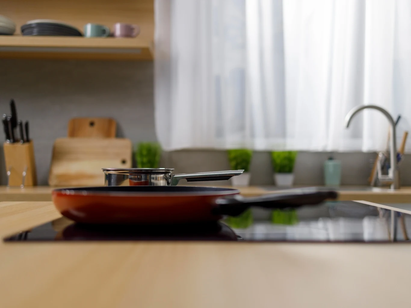 Czerwona patelnia i stalowy garnek stoją na płycie indukcyjnej na tle jasnej kuchni z drewnianym blatem, deskami do krojenia i doniczkami z ziołami przy oknie.