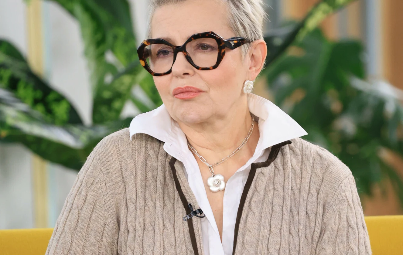 Aleksandra Konieczna Aleksandra Konieczna na żółtej kanapie, w jasnym swetrze, z siwymi włosami i okularami, na tle zielonych roślin.