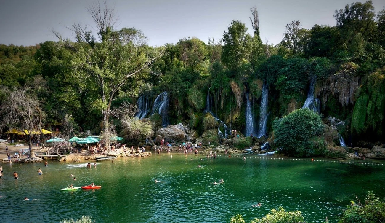 zielone jezioro otoczone bujną roślinnością, kilka wodospadów spływających ze skalistych brzegów, ludzie kąpiący się i pływający, plaża z parasolami i łódkami w tle