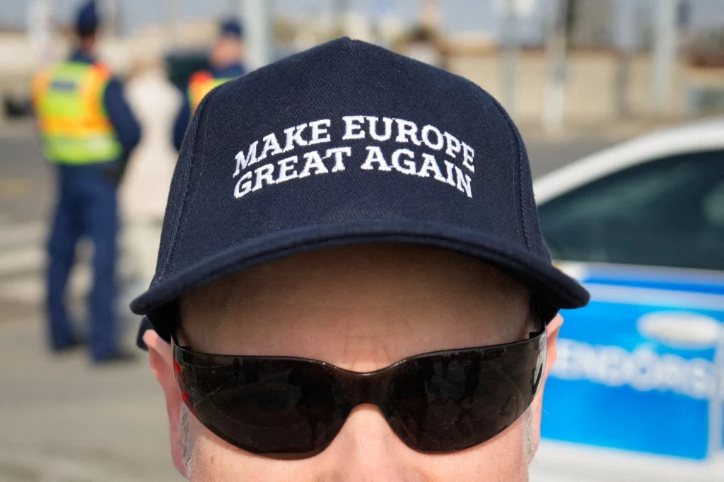 Czapka z daszkiem z napisem 'MAKE EUROPE GREAT AGAIN' na głowie osoby w ciemnych okularach przeciwsłonecznych, w tle funkcjonariusze w kamizelkach odblaskowych oraz fragment radiowozu.