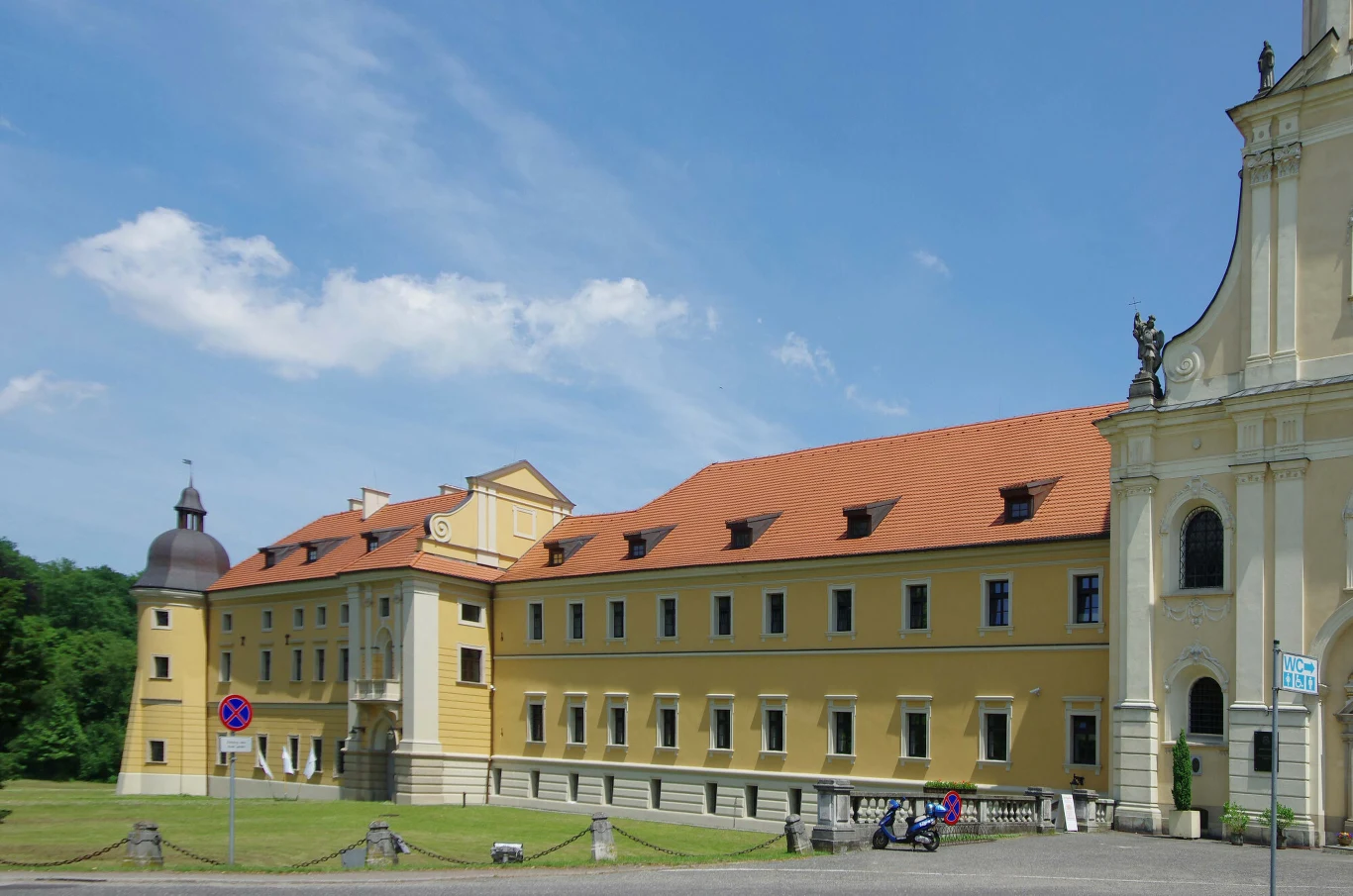 Pocysterski Zespół Klasztorno-Pałacowy w Rudach Długi, żółty budynek klasztorny z czerwonym dachem, połączony z barokowym kościołem, otoczony trawnikiem i wydzielonym chodnikiem, na tle błękitnego nieba z lekko rozproszonymi chmurami.
