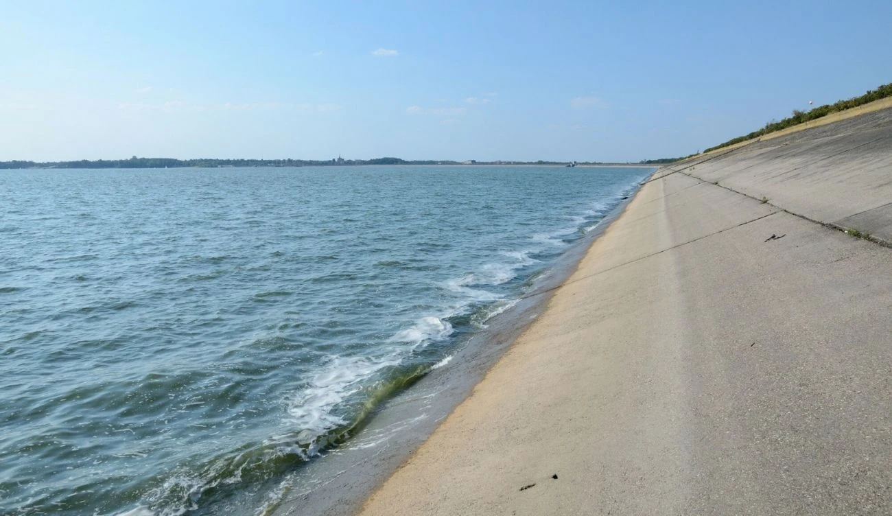 sztuczny zbiornik wodny z betonową zaporą, spokojna tafla wody sięgająca do brzegu, brak ludzi i roślinności w pobliżu brzegu, jasne niebo bez chmur