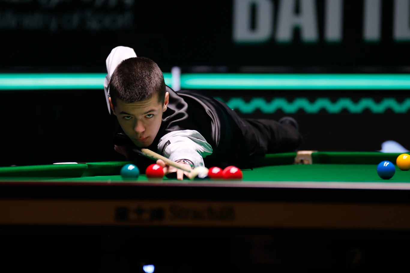 Młody mężczyzna w eleganckiej kamizelce celuje kijem bilardowym do zagrania w snooker, skupiony na uderzeniu białej bili w kierunku grupy czerwonych bil na zielonym stole.