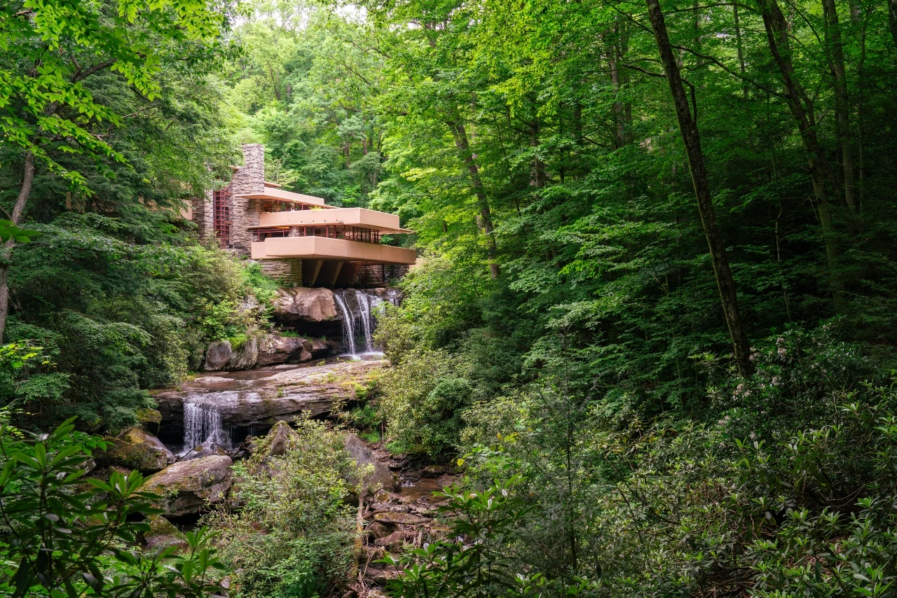 Dom nad wodospadem zaprojektowany przez Franka Lloyda Wrighta Nowoczesny dom zbudowany na skałach nad wodospadem, otoczony gęstym, zielonym lasem, z kaskadą wody spływającą bezpośrednio pod konstrukcją budynku.