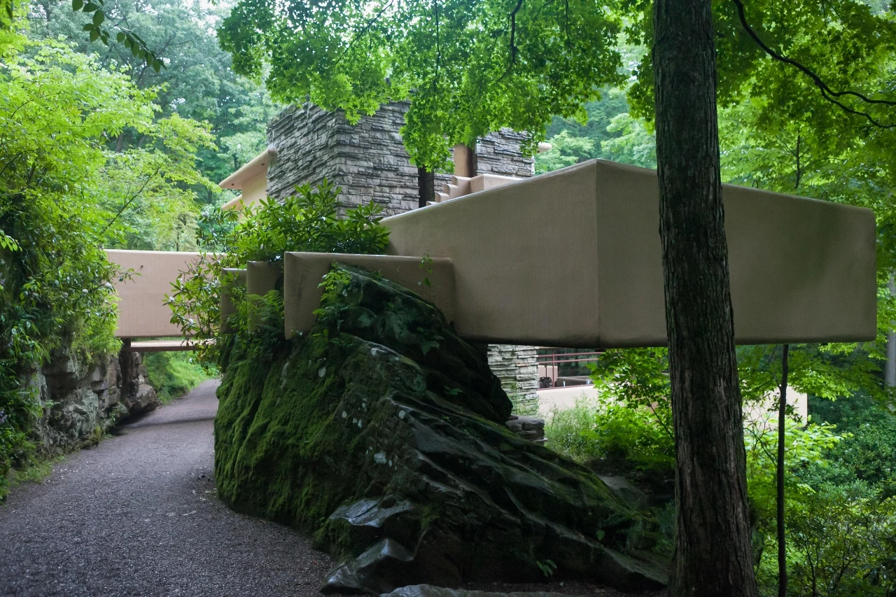 "Fallingwater", dom projektu F.L. Wrighta Nowoczesny budynek o geometrycznych kształtach harmonijnie wkomponowany w naturalny krajobraz lasu, z betonową konstrukcją przebiegającą nad dużą, omszałą skałą i otoczony bujną zielenią drzew oraz krzewów.
