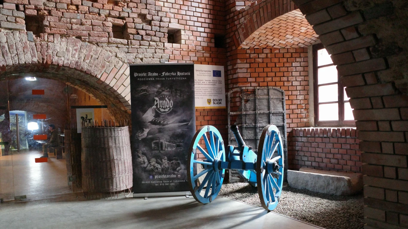 Twierdza Srebrna Góra posiadał 151a kazamat rozmieszczonych na trzech kondygnacjach. W praktyce była niemożliwa do zdobycia. niebieska armata z drewnianymi kołami ustawiona w zabytkowym wnętrzu z cegły, obok stoją czarne beczki i duży plakat informacyjny, z prawej strony światło wpada przez okno