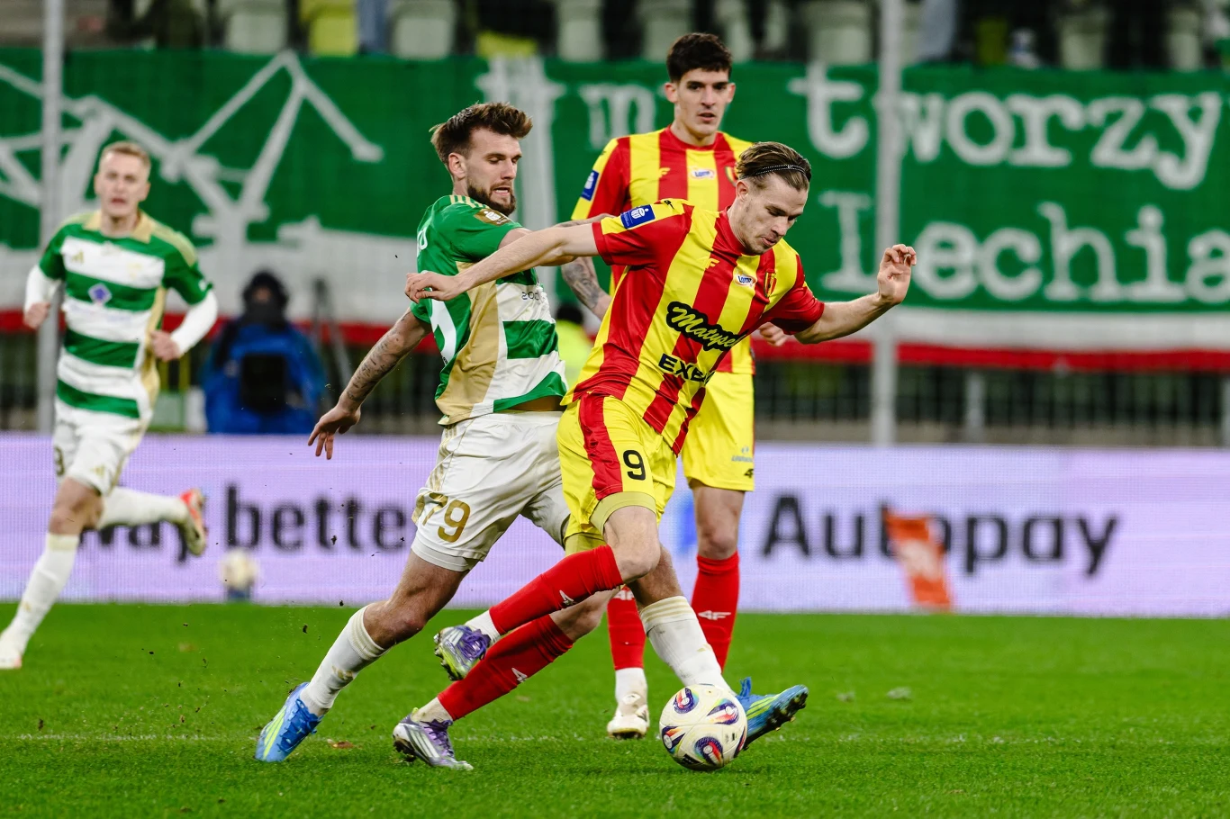Piłkarze Korony Kielce w meczu z Lechią Gdańsk dwóch piłkarzy walczy o piłkę podczas meczu, zawodnik w żółto-czerwonej koszulce próbuje ochronić futbolówkę przed rywalem w zielono-białym stroju, w tle widoczni kolejni gracze oraz zielony transparent na trybunach
