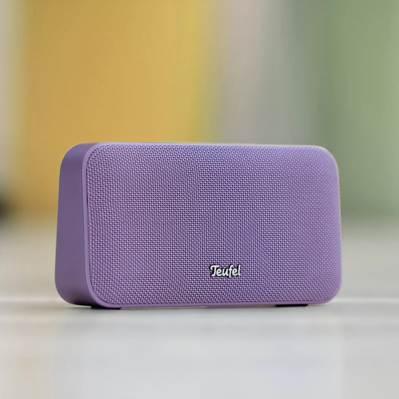 fioletowy głośnik bezprzewodowy marki Teufel z siateczkową maskownicą ustawiony na jasnej powierzchni, na tle rozmytych kolorowych pasów