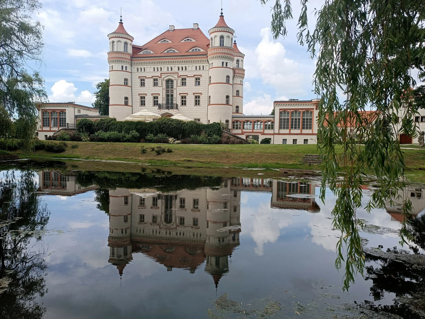 okazały pałac w stylu neorenesansowym z dwoma wieżami bocznymi i czerwoną dachówką, odbijający się w spokojnej tafli stawu otoczonej zielenią i niskimi krzewami