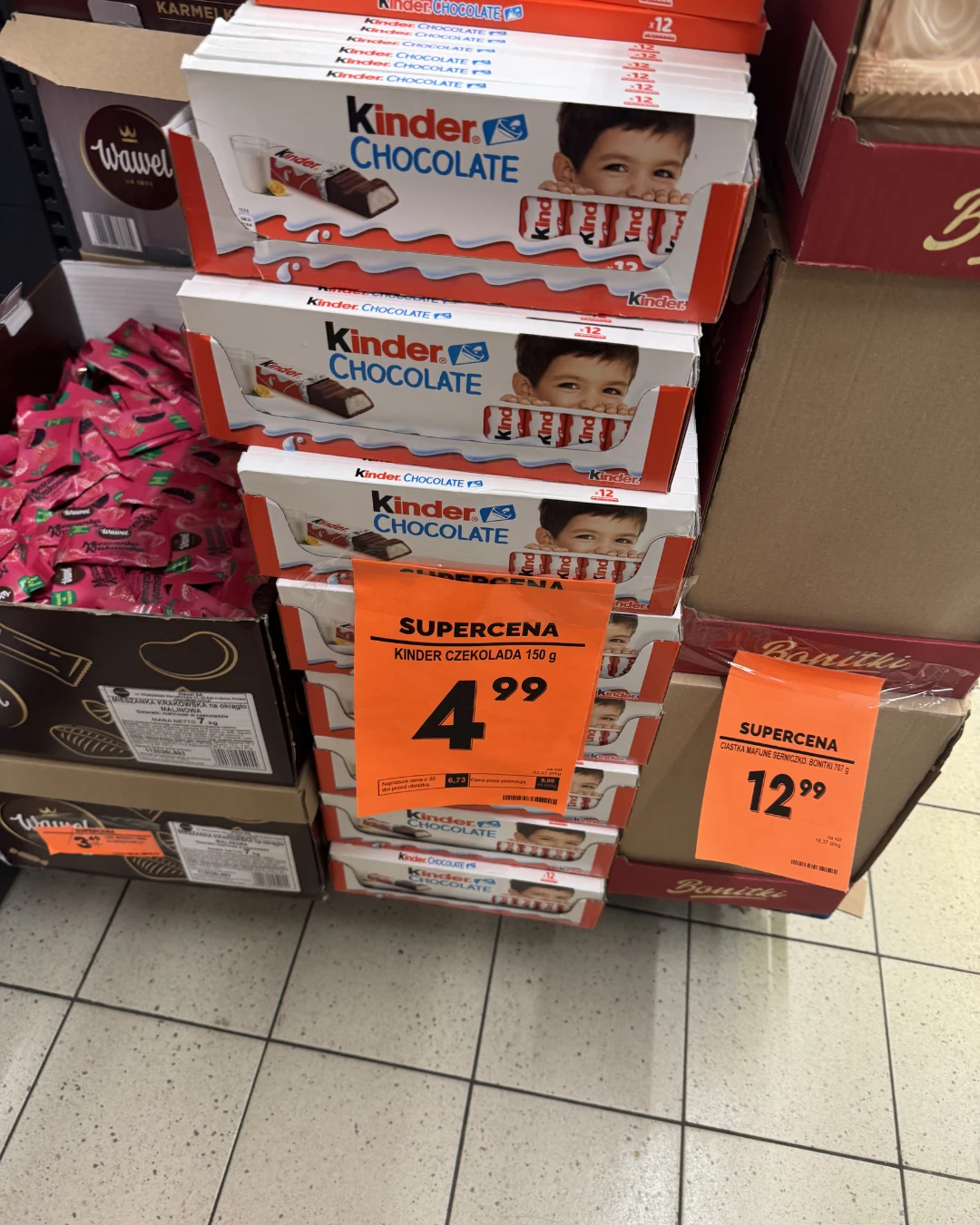 Stos opakowań Kinder Chocolate ustawionych na półce sklepowej, każde z wizerunkiem dziecka i kawałków czekolady, na środku wyraźna pomarańczowa etykieta z ceną promocyjną 4,99 zł, obok inna etykieta z ceną 12,99 zł.