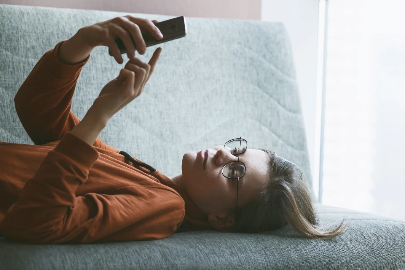 młoda osoba leżąca na kanapie, trzymająca w rękach smartfon i wpatrująca się w ekran