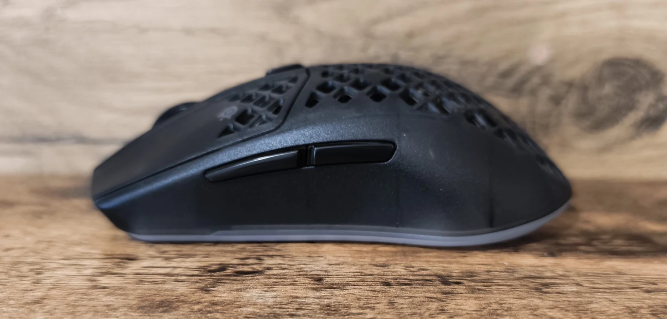 czarna mysz komputerowa z perforowaną obudową, widok z boku, dwa boczne przyciski, tło z jasnego drewna