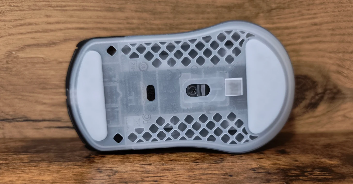 spód myszki komputerowej z przezroczystą obudową o strukturze plastra miodu, widocznymi dużymi białymi ślizgaczami oraz centralnie umieszczonym sensorem optycznym