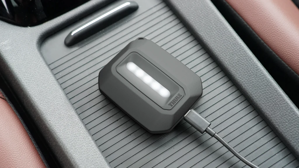 Czarny, prostokątny powerbank z podświetleniem LED podłączony kablem USB, umieszczony na gładkiej, żebrowanej powierzchni konsoli samochodu z widoczną brązową tapicerką siedzenia po bokach.