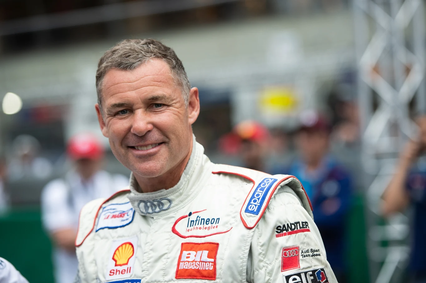 mężczyzna w kombinezonie wyścigowym z licznymi sponsorami, uśmiechnięty, stoi na tle rozmytego tłumu osób i elementów toru wyścigowego