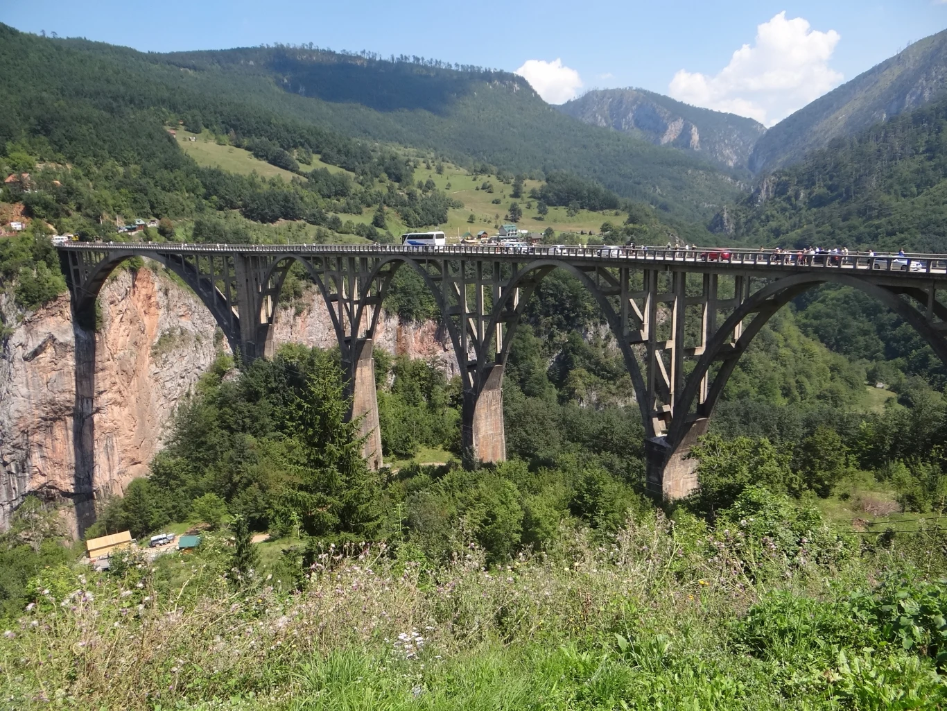 masywny betonowy most łukowy rozpięty nad głębokim, zielonym wąwozem, po którym poruszają się samochody, a w tle rozciągają się gęsto zalesione wzgórza