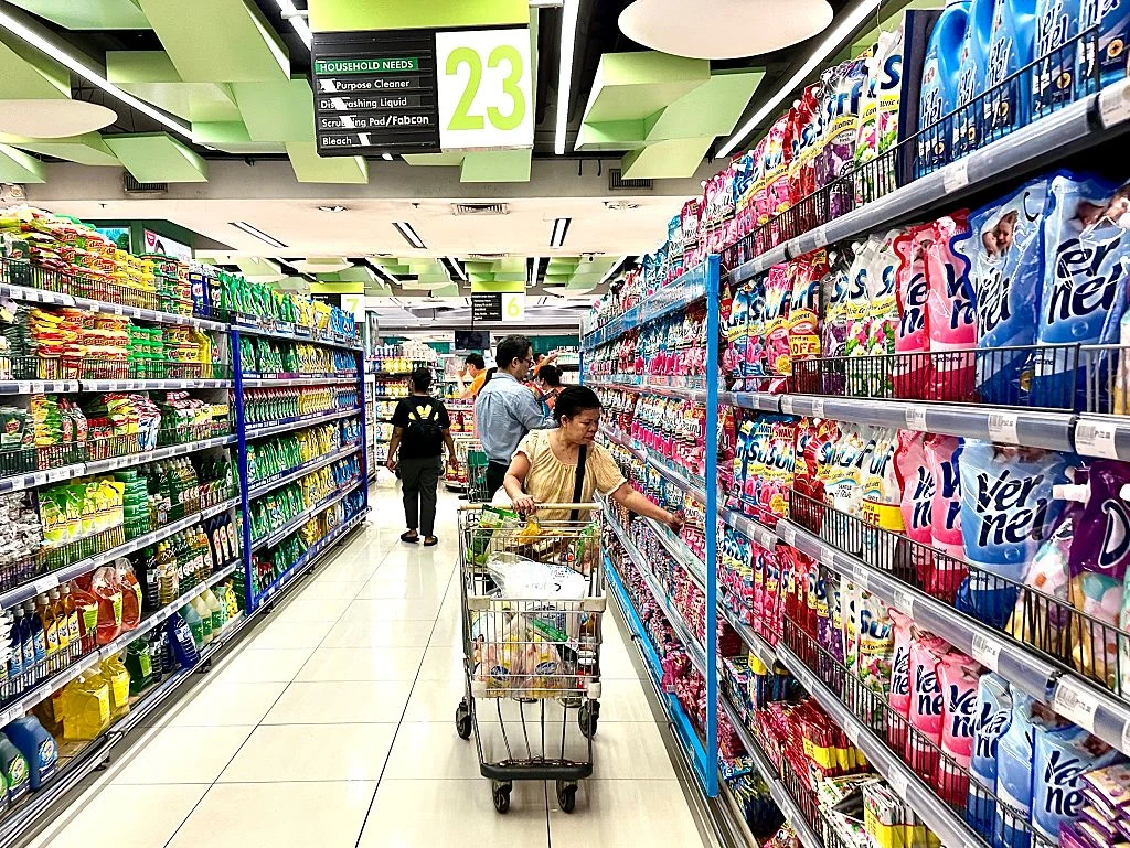 kobieta pcha wózek sklepowy w szerokiej alejce supermarketu, po lewej stronie regały z produktami spożywczymi, po prawej strony kolorowe opakowania płynów do płukania tkanin, w tle kilku innych klientów