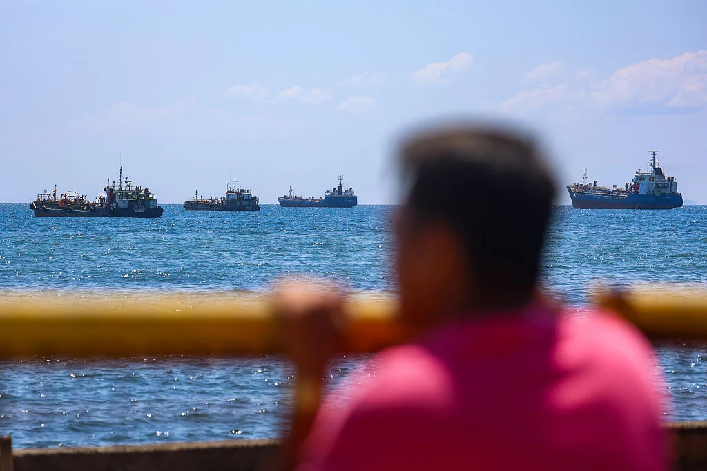 Tankowce z surowcami u wybrzeży Filipin Kilka dużych statków płynie po spokojnym morzu, w centrum uwagi rozmyta sylwetka osoby w różowej koszulce opierającej się o żółtą barierkę na pierwszym planie, błękitne niebo z lekkimi chmurami w tle.