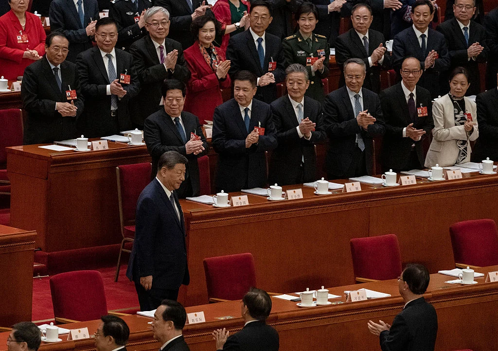 Na kryzysie bliskowschodnim ewidentnie skorzystały Chiny Mężczyzna w garniturze przechodzi między rzędami czerwonych foteli, wokół niego stojący i siedzący urzędnicy i politycy w garniturach klaszczą, większość osób nosi identyfikatory przypięte do klap marynarek, w tle ciemne biurka z filiżankami.