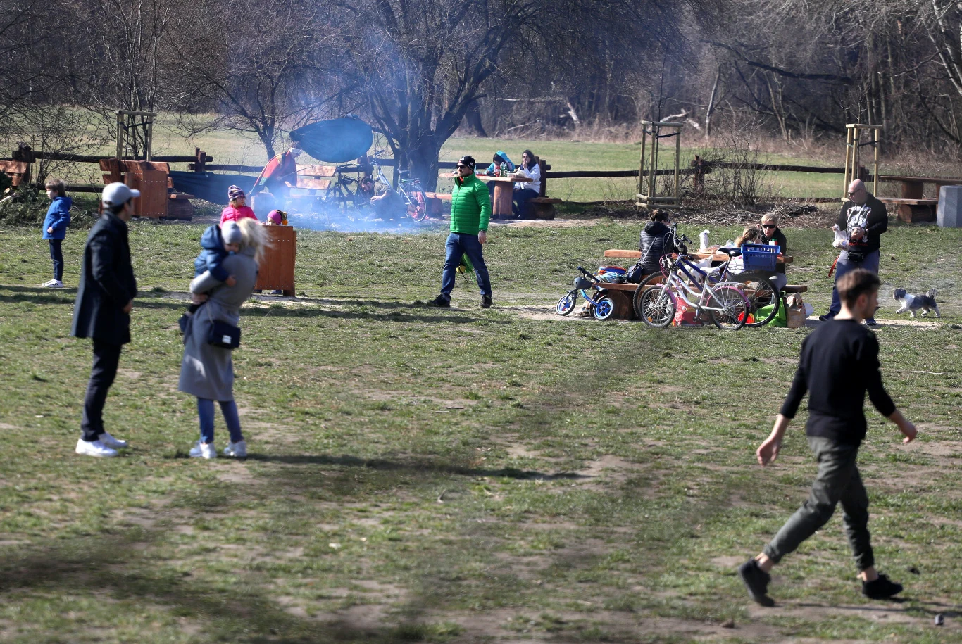 Kilka osób spaceruje i spędza czas na świeżym powietrzu w parku, w tle widać grupę przy ognisku oraz rowery rozstawione na trawie.