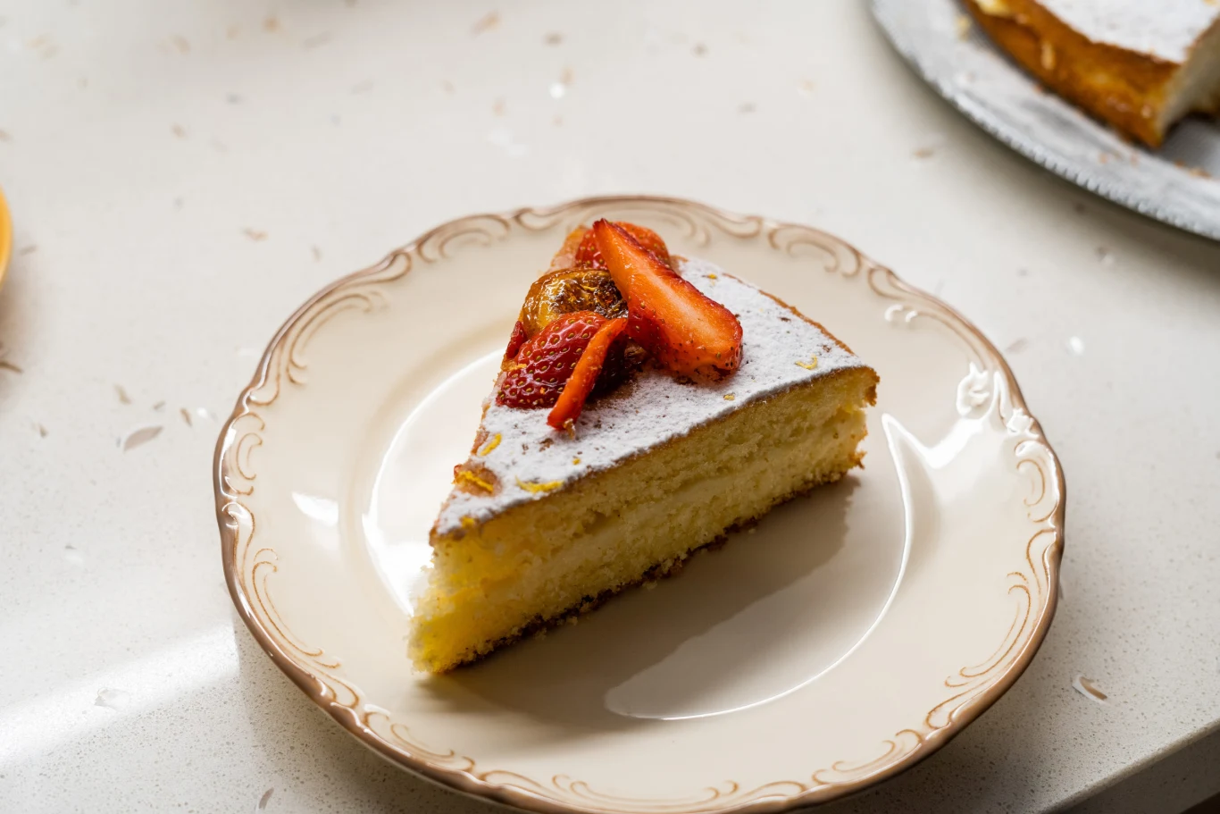 Kawałek jasnego biszkoptowego ciasta oprószonego cukrem pudrem i udekorowanego plasterkami truskawek, podany na eleganckim talerzyku z delikatnym wzorem na brzegu.