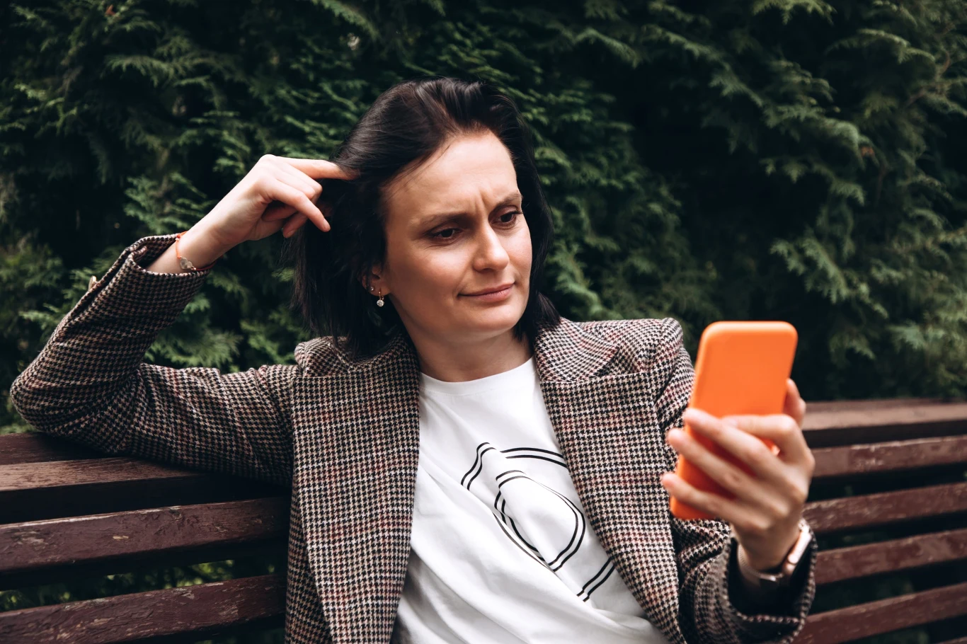Kobieta w marynarce w kratę i białej koszulce siedzi na ławce, trzymając w dłoni pomarańczowy telefon i patrząc na ekran z lekkim wyrazem konsternacji na twarzy, w tle zielone liście drzew.