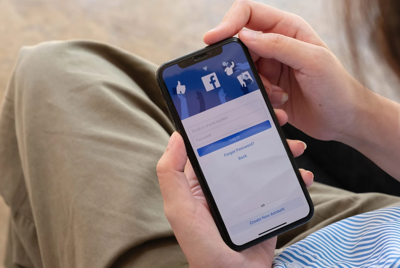 osoba trzyma w dłoniach smartfona z włączonym ekranem logowania do aplikacji Facebook, na którym widoczne są opcje wpisania hasła oraz logowania