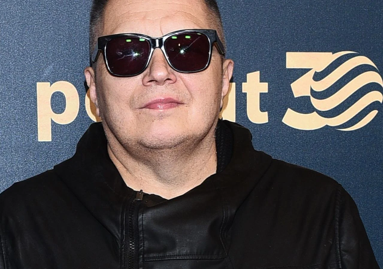 Muniek Staszczyk Muniek Staszczyk w ciemnych okularach i czarnej kurtce na tle granatowej ścianki z żółtymi napisami.