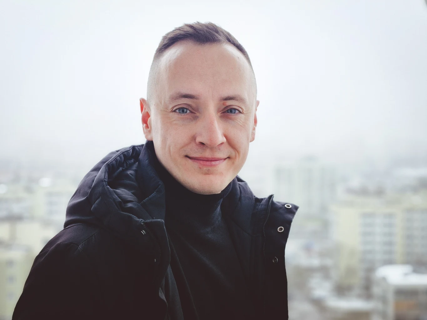 Mężczyzna z krótkimi włosami ubrany w czarną kurtkę, stojący na tle zamglonego miasta z widocznymi blokami w oddali.