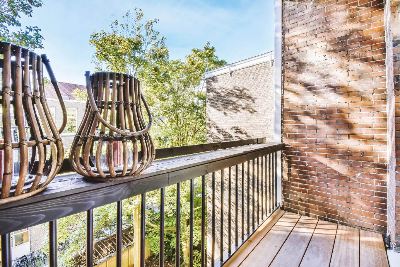 Drewniany balkon z metalową balustradą, na którym stoją dwa dekoracyjne lampiony z wikliny; w tle widoczne są drzewa i ceglana ściana sąsiedniego budynku.