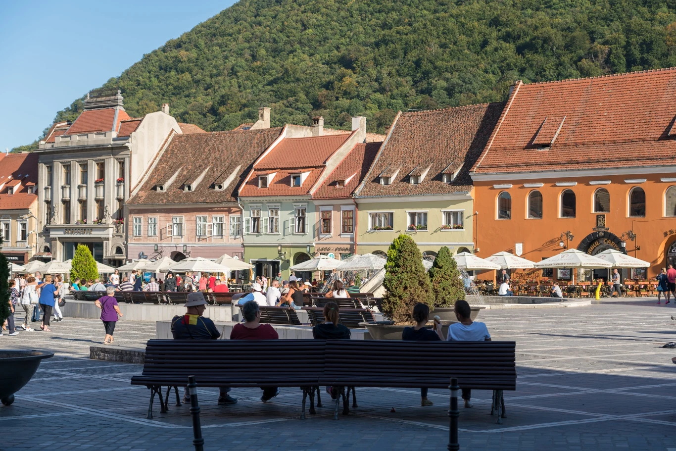 Rząd kolorowych, zabytkowych kamienic z czerwonymi dachami otacza przestronny plac miejski, na którym ludzie siedzą na ławkach i spacerują, a w tle widoczne są zielone zbocza wzgórza pokrytego drzewami.